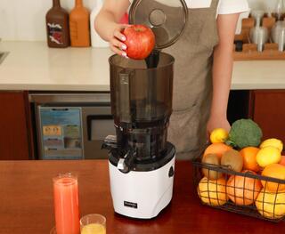 Foto einer Person in der Küche mit einem Korb voller Obst und Gemüse, die einen ganzen Apfel in den Kuvings AUTO10 Slow Juicer legt.